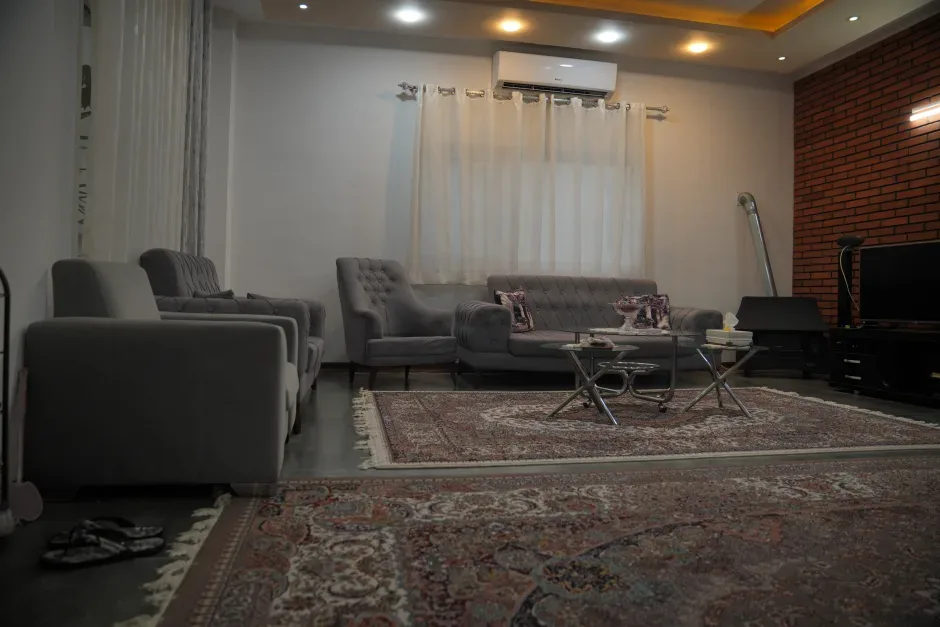 پذیرایی مبله‌ی یک ویلا: مبل‌های طوسی، فرش ایرانی، دیوار آجری و پرده‌ی سفید